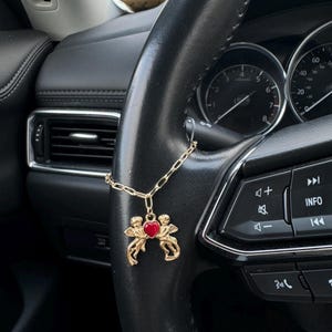 May include: A gold charm depicting two cherubs holding a red heart, suspended from a gold chain. The charm hangs from a black steering wheel with various buttons and dials. The steering wheel is in a car.