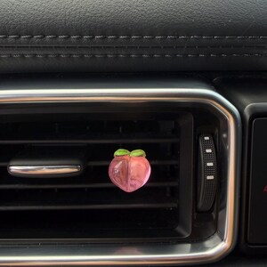 以下が含まれることがあります： 緑の葉のアクセントが付いた、ピンク色の桃の形をした車の芳香剤。芳香剤は、シルバーのトリムが付いた黒い車の通気口に取り付けられています。車の内装は黒革です。