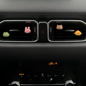 May include: A car dashboard featuring four cartoon air fresheners. The air fresheners are designed as a green frog, a pink bunny, a brown bear, and a yellow chick. They are positioned in the air vents of the car.
