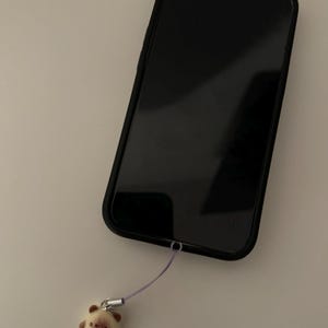 May include: Black smartphone in a black case with a small, beige bear charm attached by a purple cord. The bear charm has brown accents and a silver-colored clasp.