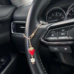 May include: A gold-colored car charm hanging from a steering wheel. The charm features a red strawberry with green leaves and a white daisy. The steering wheel is black, and the car's interior is visible in the background.