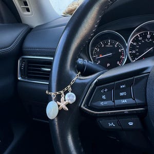 Puede incluir: Un dije de coche dorado con una cadena, con un óvalo blanco, una estrella de mar y un dije redondo. El dije cuelga del volante de un coche, con el salpicadero y los indicadores visibles en el fondo.