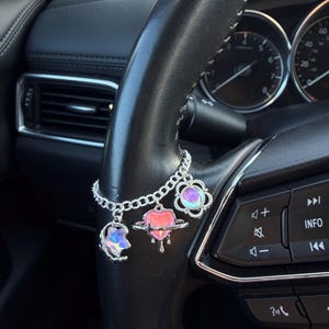 May include: A silver charm bracelet with three celestial-themed charms hanging from a car steering wheel. The charms include a crescent moon, a heart with a planet design, and a flower. The charms have iridescent and pink accents.