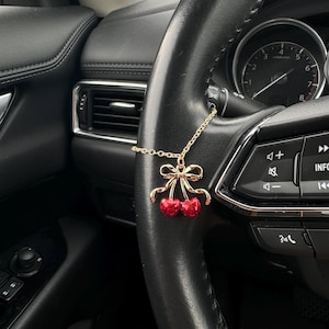 May include: A gold-colored cherry charm with red cherries and a bow, hanging from a car's steering wheel. The steering wheel is black, and the car's interior is visible in the background.