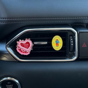 May include: Two enamel pins are displayed on a car's air vent. One is heart-shaped with the text "Literally just the best" and the other is a yellow circle with a Puerto Rico design and the text "Seguimos Aqui".