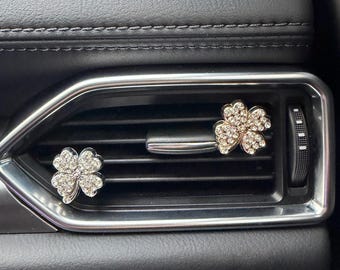 May include: Two four-leaf clover shaped car air vent clips decorated with clear rhinestones. One clip has a silver-tone metal finish, while the other has a gold-tone metal finish. Both ornaments are attached to the horizontal slats of a vehicle air vent.