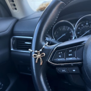 May include: A gold bow charm on a pearl necklace hangs from a black steering wheel. The bow is the focal point. The steering wheel has buttons and dials, and is in a car.