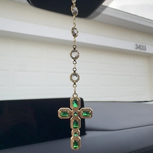 May include: Gold-toned car charm featuring a cross pendant with emerald-green gemstones. The charm hangs from a chain of clear, faceted beads and is suspended from a car's rearview mirror.