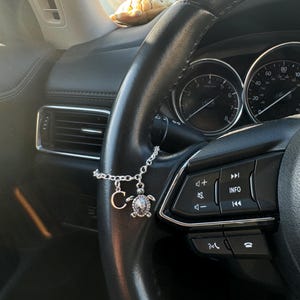 May include: A silver car charm with a turtle pendant and the letter "C" hanging from a steering wheel. The charm is attached to the steering wheel with a silver chain. A plush turtle is on the dashboard.