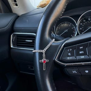 May include: A silver car charm featuring a cross pendant with a red heart-shaped gem in the center. The charm hangs from a chain attached to a car's steering wheel. The steering wheel is black leather.