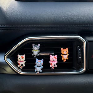 May include: Six colourful cat-shaped car vent clips. The cats have various patterns and colours, including orange, pink, blue, and grey. They are attached to the car's air vent, adding a decorative touch to the interior.
