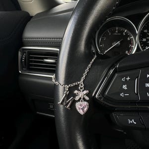 May include: A silver car charm suspended from a black steering wheel. The charm includes the letter "M", a bow, and a pink heart-shaped pendant. The car's interior is visible in the background.