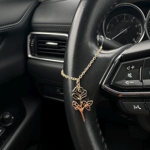 May include: A gold-colored rose-shaped car charm hanging from a gold chain. The charm is suspended from a black steering wheel, with a dashboard and other car features visible in the background.