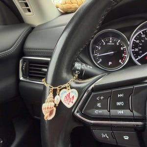 May include: A gold-colored charm hanging from a car's steering wheel. The charm features a seashell, a pearl, and a heart-shaped pendant with a red fish design. The steering wheel is black.