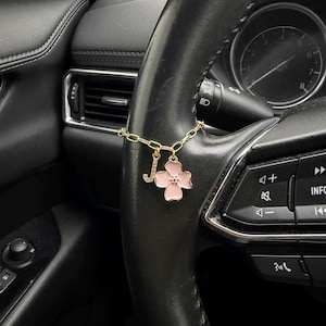 May include: A gold-toned chain charm featuring a letter J pendant encrusted with small clear stones and a four-petaled flower charm with light pink enamel, attached to a black leather-wrapped steering wheel in a vehicle interior.