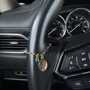 May include: Gold car charm suspended from a black steering wheel. The charm includes a small blue gem and an oval pendant with a floral design. The car's dashboard and gauges are visible in the background.
