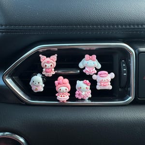 May include: Six pink and white cartoon character air vent charms are displayed. The charms, featuring bows and other decorations, are attached to a car's air vent. The dashboard is dark grey.