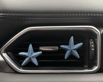 May include: Two textured, blue starfish-shaped car air vent clips attached to the horizontal slats of a vehicle's rectangular air conditioning vent, which features a silver metallic border and a black plastic housing.