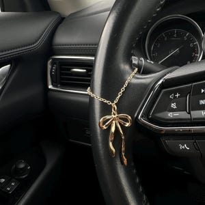 May include: A gold bow-shaped car charm suspended from a black steering wheel. The charm hangs by a fine chain. The steering wheel has integrated controls. The car's interior is predominantly black, with visible air vents and dashboard.