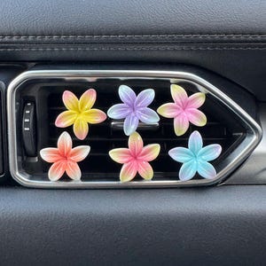 May include: Six flower-shaped car vent clips in various colours. Each clip has a gradient effect, with shades of yellow, orange, pink, purple, and blue. They are placed in a car vent, against a black dashboard.