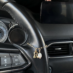 Op de afbeelding: Een goudkleurige kettingarmband met een letter R-bedel en een witte ovale parelbedel, gewikkeld om het zwarte lederen stuurwiel van een voertuig. De bedel bevindt zich bij de dashboardbediening en een klein figuurtje op het display.