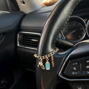May include: Gold-colored car charm with a baby-themed design. The charm features a teddy bear, a baby bottle with a heart, and the word "MAMA". The charm hangs from a car's steering wheel.