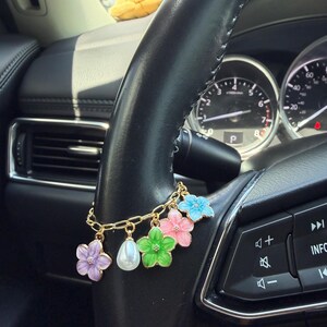 May include: A gold-toned chain bracelet featuring five dangling charms: a purple flower, a white teardrop-shaped pearl, a green flower, a pink flower, and a light blue flower. The bracelet is wrapped around the black leather steering wheel of a vehicle.
