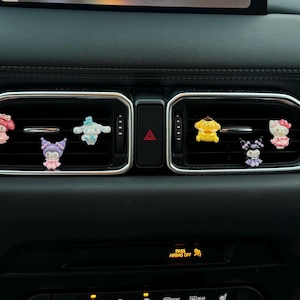 May include: A car dashboard featuring six cartoon character air vent decorations. The characters include a pink Hello Kitty, a blue Cinnamoroll, a yellow Pompompurin, and others in purple and pink. The dashboard is black.