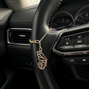 May include: A gold-colored butterfly car charm hanging from a black steering wheel. The charm is a detailed, openwork design of a butterfly wing. The car's dashboard and controls are visible in the background.