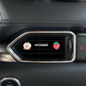 May include: Two car vent charms: a white cupcake with red detailing and a red strawberry with green leaves. The charms are attached to a car vent, adding a decorative touch to the vehicle's interior.