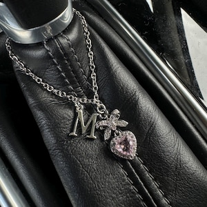 May include: Silver necklace with an initial "M" pendant, a bow, and a pink heart-shaped charm. The necklace is displayed on a black leather surface, showcasing the jewelry's details and design.