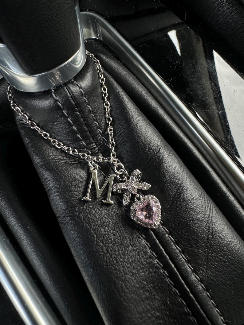 May include: Silver necklace with an initial "M" pendant, a bow, and a pink heart-shaped charm. The necklace is displayed on a black leather surface, showcasing the jewelry's details and design.