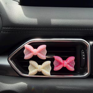 May include: Three decorative car air vent bow accessories. The bows are in shades of pink and white, and are covered in small, sparkling embellishments. The bows are placed in a car's air vent.