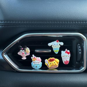 May include: Six colourful car vent charms, each featuring cartoon characters and dessert designs. The charms include a cupcake, ice cream cone, and a milkshake. The glossy charms are designed to clip onto a car's air vent, adding a decorative touch.