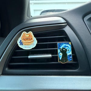 May include: Two car air vent decorations. One is a cat-shaped pancake design on a blue plate. The other is a black cat against a starry night background, reminiscent of Van Gogh's style.