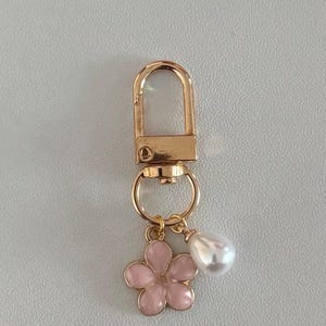 May include: A gold-coloured keychain with a lobster clasp. It features a pink flower charm and a white teardrop-shaped pearl. The flower has five petals and a gold outline. The keychain is designed for attaching to keys or bags.