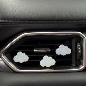 May include: Three cloud-shaped car air fresheners are placed in a car's air vent. The air fresheners are white and have a textured appearance. The car's interior features a black dashboard and silver air vent trim.