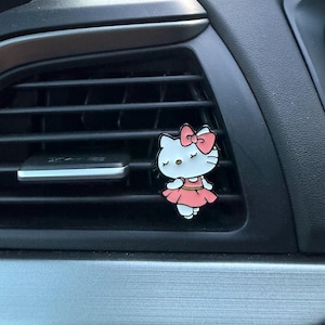 May include: A Hello Kitty car air vent clip. The clip features a white cat with a pink bow, dress, and accents. The clip is attached to a black car air vent.