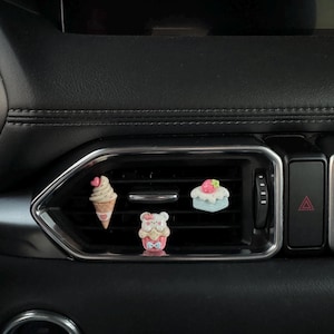 May include: Three decorative car vent clips. One is an ice cream cone with a pink heart, another is a bear with a bow, and the last is a cake with a strawberry on top. The clips are in a car vent.