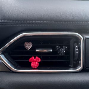 May include: Three car air vent decorations: a silver glitter heart, a black glitter Mickey Mouse head, and a red glitter Minnie Mouse bow. The decorations are placed in a car's air vent.