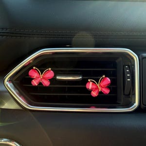May include: Two small, translucent pink butterfly-shaped car air freshener clips with gold-toned wire frames. Each butterfly features a vibrant pink hue and is attached to the horizontal slats of a black vehicle air conditioning vent.