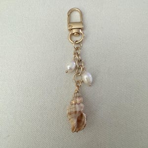 May include: A gold-tone keychain featuring a lobster clasp, chain, and three white pearls. A small, tan seashell with gold detailing dangles from the chain. The keychain is designed for keys or as a decorative accessory.