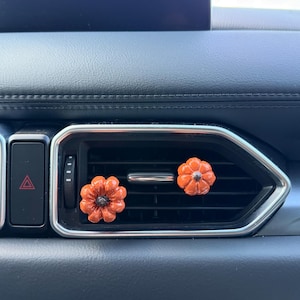 May include: Two small, glossy orange pumpkin-shaped car air freshener clips attached to the horizontal slats of a black vehicle air conditioning vent. Each pumpkin features a dark brown stem at the center and a smooth, reflective surface finish.