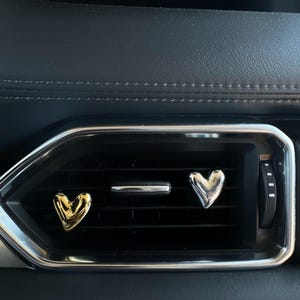 May include: Two heart-shaped car vent air fresheners. One is gold, the other silver. They are placed in a black car vent with a silver trim. The background is a black car dashboard.