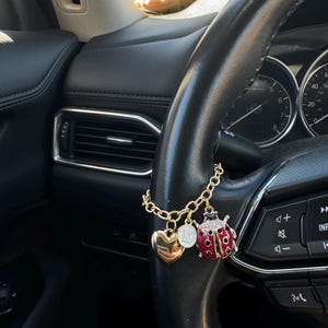 Puede incluir: Un charm de coche dorado con un corazón, un disco con la letra "K" y un charm de mariquita roja con lunares negros. El charm cuelga de una cadena dorada sujeta a un volante negro.