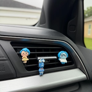 May include: Three colourful car air vent decorations featuring characters from the animated film "Inside Out." The figures are Joy, Sadness, and Anger, each with distinct facial expressions and outfits. The decorations are attached to the car's air vent.