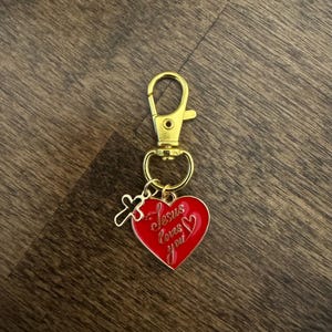 May include: A gold-coloured keychain with a lobster clasp. The keychain features a red heart pendant with the words "Jesus loves you" in cursive script. A small gold-coloured cross charm hangs next to the heart.