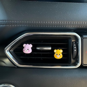 May include: Two cartoon bear-shaped car air vent decorations. One is pink, the other yellow. Both have smiling faces and are placed on a black car vent with a silver trim.