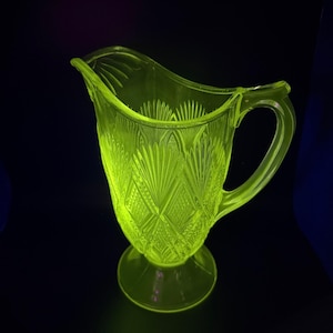 Op de afbeelding: Een gloeiende glazen kan in vintage-stijl met een handvat en een tuit. De kan heeft een decoratief patroon van ruitvormen en waaiervormige ontwerpen. Het glas heeft een heldere, neon groene kleur en staat op een ronde voet.