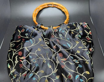 Bolso de mano negro de satén bordado con flores, asas circulares de bambú y cierre de cremallera superior.